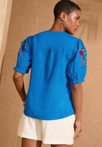 Blusa blu con texture e maniche a sbuffo, caratterizzata da ricami floreali colorati sulle spalle e un design con schiena leggermente arricciata.