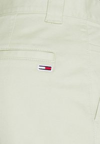 Pantaloni di cotone verde chiaro con una texture liscia, dotati di un piccolo logo ricamato in rosso, bianco e blu sulla tasca posteriore.