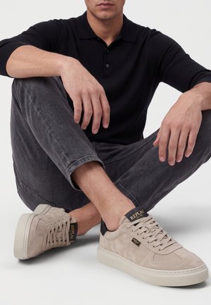 Homme portant un polo noir à manches longues, un jean gris foncé et des baskets beiges Replay, assis en tailleur sur un sol blanc.