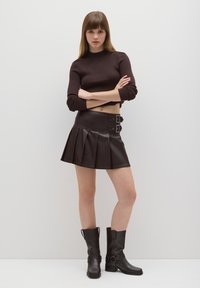 Top corto marrone a coste, minigonna plissettata in pelle scura con fibbie decorative e stivali neri al ginocchio in pelle con punta quadrata.