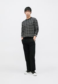 Juodas megztinis su geometriniu raštu, dėvimas su juodomis kelnėmis ir juodais sportbačiais, megztinio rankogaliai su baltais akcentais.