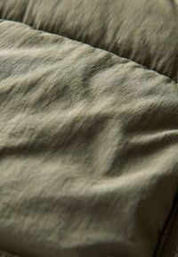 Tejido texturizado en verde oliva con una superficie suave y ligeramente arrugada. El material parece ligero y duradero, con detalles de costura visibles.