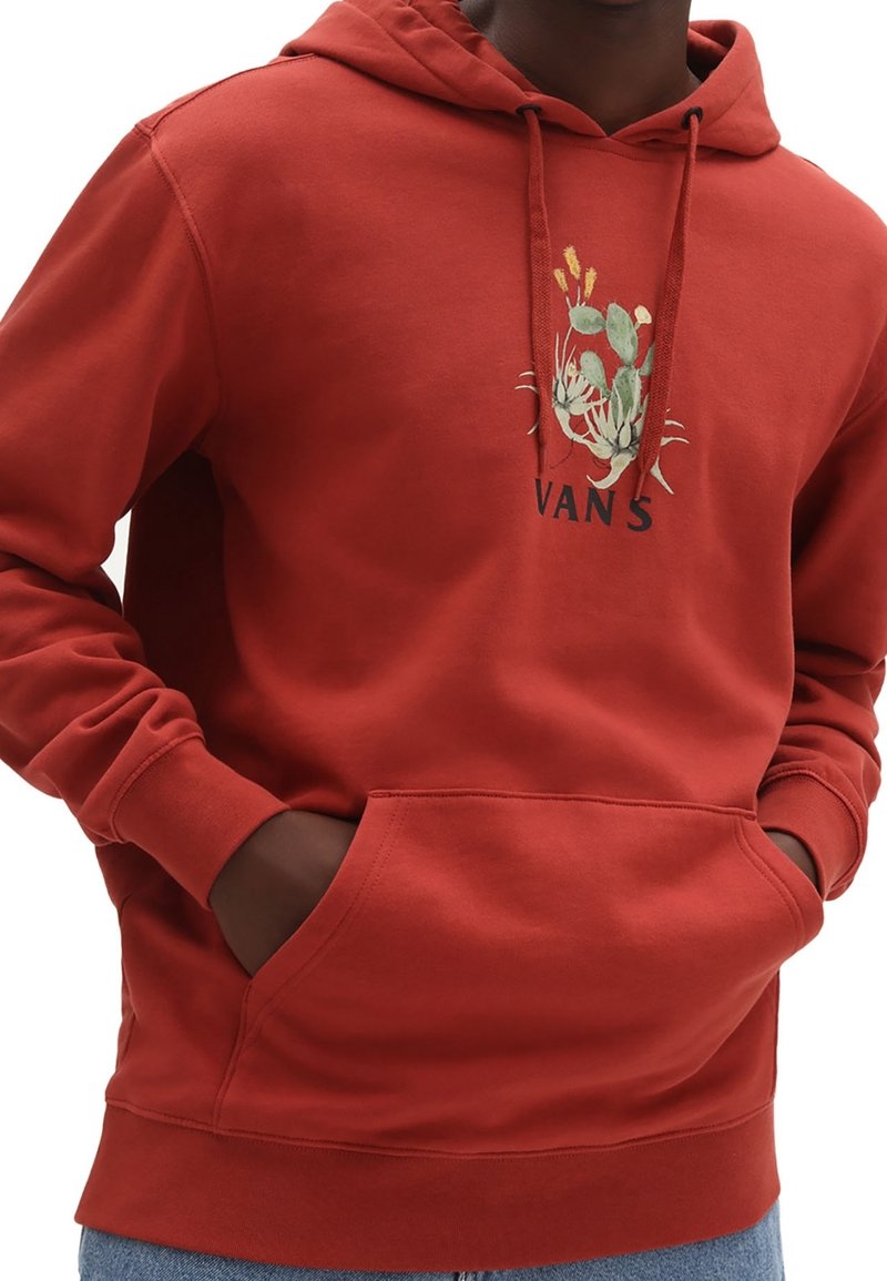 Homme portant un sweat à capuche Vans couleur rouille avec un motif cactus et une poche avant, mains partiellement glissées à l'intérieur, jean bleu visible.