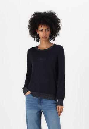 Maglione navy con scollo rotondo, polsini e orlo in maglia testurizzata. Dettaglio logo discreto sul petto. Abbinato a jeans azzurri chiari.