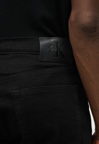 Κοντινό πλάνο ενός ατόμου που φοράει μαύρα τζιν με μαύρο δερμάτινο ταμπελάκι με το λογότυπο Calvin Klein πάνω από την πίσω τσέπη.