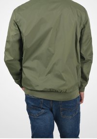 Chaqueta bomber verde oliva con un acabado suave y brillante, y puños y dobladillo acanalados. Combinada con jeans de mezclilla azul que presentan bolsillos traseros con costuras.