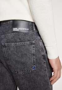 Fekete farmernadrág texturált felülettel és kék díszvarrással. Fekete címke található rajta, fehér betűkkel: "KARL LAGERFELD JEANS".