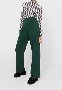 Grüne weite Hosen mit hoher Taille, ausgestattet mit Vorderfalten und einem Knopfverschluss. Kombiniert mit einem gemusterten langärmeligen Oberteil und klobigen Schuhen.