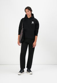 Sweatshirt preto com logótipo, bolso frontal e cordões, combinado com calças pretas e sapatilhas altas pretas. Mala de ombro cinza.