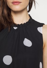 Svart ärmlös klänning med en volangkrage och stora vita prickar. Tyget verkar lätt och mjukt.