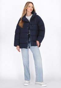 Piumino navy con colletto alto, chiusura a bottoni e texture trapuntata orizzontale. Abbinato a jeans azzurri chiari e scarpe bianche.