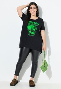 Black oversized t-shirt with green "DISCO NIGHT CLUB" graphic, paired with shiny black leggings and leopard-print slip-on shoes, holding a green bag.