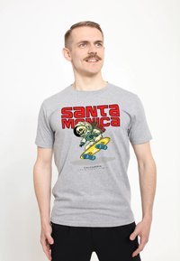 Muž s knírem, oblečený v šedém tričku s potiskem skatebordingového astronauta s kostrou a červeným textem "Santa Monica".