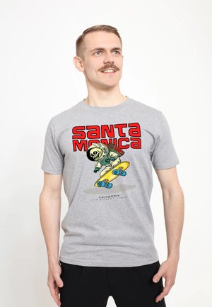 Muž s knírem, oblečený v šedém tričku s potiskem skatebordingového astronauta s kostrou a červeným textem "Santa Monica".
