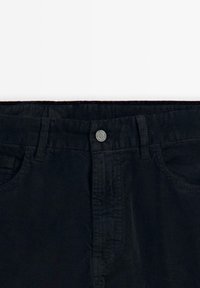 Svart kordfløyelsbyxor med en slät textur, utrustad med metallknappstängning och två framfickor. Synlig vertikal ribbad struktur längs tyget.