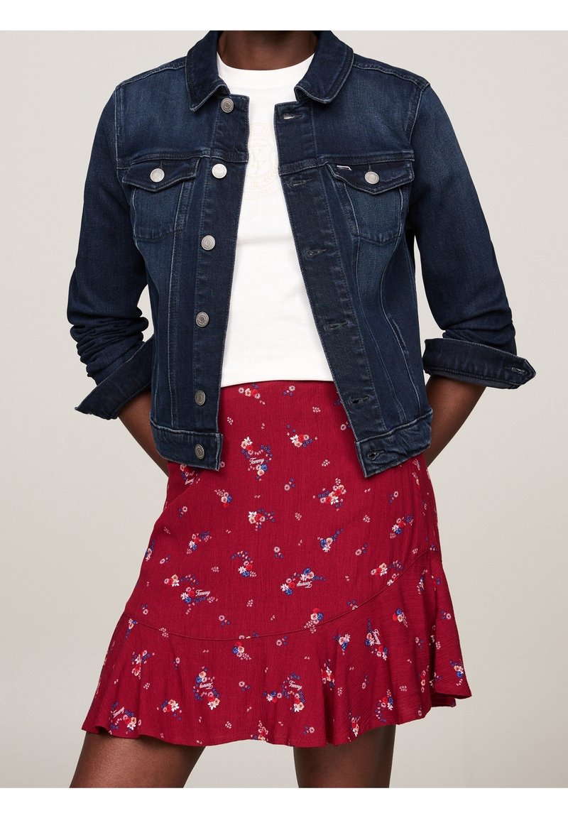 Žena v tmavě modré džínové bundě přes bílé tričko a červené květinové mini sukni, stojí s rukama v kapsách.