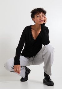 Cardigan noir avec dentelle, manches côtelées et boutons, associé à un pantalon gris ample et des bottes plateforme noires, modèle accroupi contre un fond blanc.