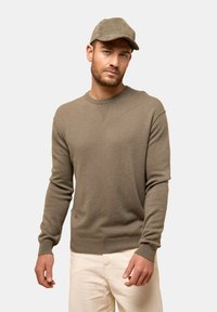 Brun stickad tröja med rund halsringning, ribbade ärmslut och nederkant, kombinerad med krämfärgade sammetsbyxor och en matchande brun keps.