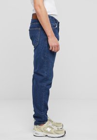Jeans de mezclilla azul oscuro con corte ajustado, parche de cuero marrón en la cinturilla y una vista lateral que muestra la textura y el contorno de la tela.