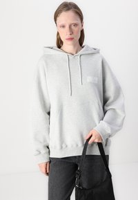 Grå hettegenser laget av mykt stoff, med lommer foran, snøring på hetten, og en liten logo på venstre bryst. Kombinert med mørke jeans.
