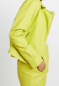 Kožená motorkářská bunda v limetkově zelené barvě s přiléhavým střihem, zapínáním na zip a bočními kapsami, s hladkou texturou a minimalistickými detaily.