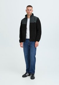 Giacca in pile nera con yoke grigio scuro, colletto alto; t-shirt bianca sotto, jeans blu, scarpe nere con strisce bianche.