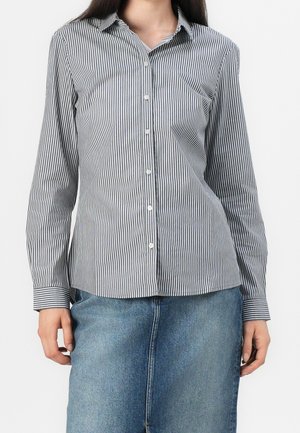 Femme portant une chemise à manches longues à rayures verticales noires et blanches avec une jupe en jean bleue, les bras détendus le long du corps.
