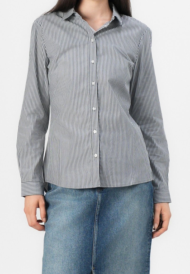 Femme portant une chemise à manches longues à rayures verticales noires et blanches avec une jupe en jean bleue, les bras détendus le long du corps.