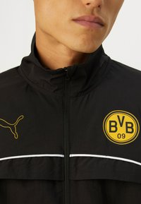 Sort atletisk jakke med lynlås, der har et gult Puma-logo og et rundt gult BVB 09 mærke. Hvide detaljer langs brystet. Teksturstof.