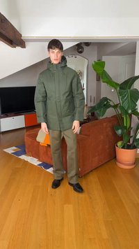 Młody mężczyzna w zielonej kurtce z kapturem, brązowych spodniach i czarnych butach stoi w salonie z drewnianą podłogą i dużą doniczką z rośliną.