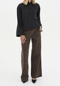 Camisa negra de manga larga con cuello y detalle de botones, combinada con pantalones sueltos marrones y tacones negros de punta. Textura de tela suave.