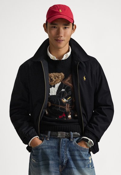 Black jacket, white shirt collar, black sweater with a bear graphic in a tuxedo, denim jeans, red cap, and a silver watch. Casual attire.
