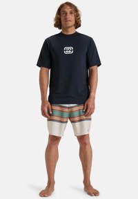 Marinblå kortärmad rash guard med logotyp, kombinerad med beiga surfshorts med horisontella ränder i orange, grönt och blått.