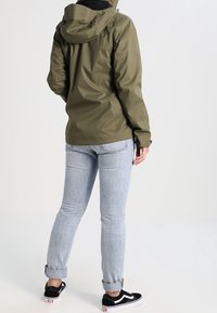 Olivengrüne wasserdichte Jacke mit Kapuze, die eine glatte Textur aufweist, kombiniert mit gerollten hellblauen Jeans und schwarzen Sneakers.