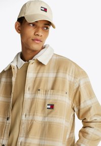 Camisa de franela a cuadros beige con cuello de sherpa blanco, bolsillo en el pecho que presenta un parche de logo, llevada sobre un sudadera ligera beige.