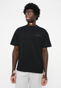 Sort kortærmet t-shirt med rund hals, prydet med et mat sort "ellesse" logo på brystet, kombineret med grå sweatpants.