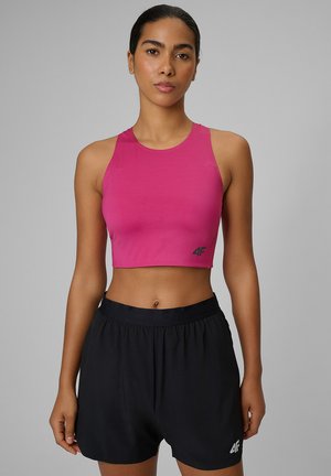 Kobieta z ciemnymi włosami związanymi do tyłu, ubrana w jasnoróżowy, bezrękawnikowy crop top i czarne sportowe szorty, stojąca na tle szarego tła.
