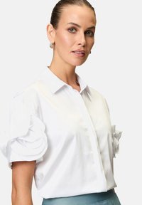 Camicia bianca a bottone con maniche puff e dettagli a volant a strati. Tessuto liscio con colletto strutturato, abbinata a una cintura di colore azzurro chiaro.