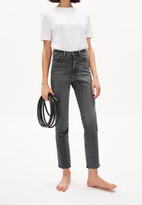 Vaqueros grises de corte ajustado y talle alto, con cinco bolsillos, sutiles desgastes y dobladillo sin rematar, combinados con una camiseta blanca sencilla y sosteniendo un cinturón negro.