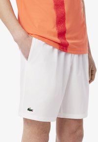 Shorts sportivi bianchi con tasche laterali e un logo Lacoste verde ricamato sull'orlo inferiore. Realizzati in tessuto leggero e traspirante.
