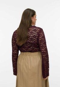 Bordeauxrode kanten top met lange mouwen gedragen over een beige rok met een elastische tailleband. Het kant heeft bloemmotieven en een doorschijnende textuur.