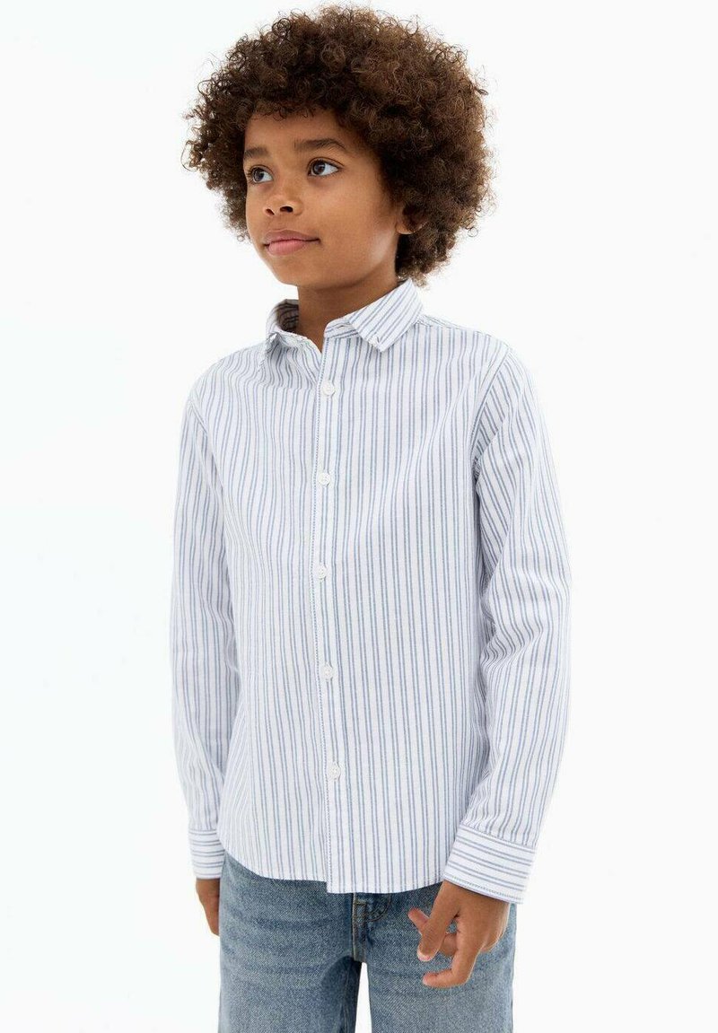 Cămașă cu dungi în albastru deschis și alb, având guler deschis și mâneci lungi, purtată cu blugi din denim albastru.