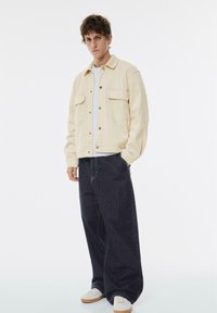Cremefarvet jakke med to brystlommer og knapper, parret med løse mørke denimbukser og hvide sneakers. Ensfarvet baggrund.