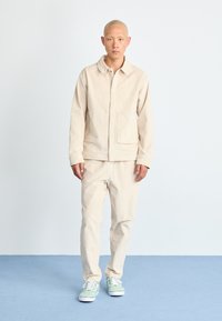 Chaqueta beige ligera con bolsillos frontales y cuello, combinada con pantalones beige a juego y zapatillas de lona verdes sobre una superficie azul.