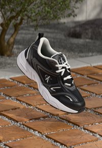 Μαύρα και άσπρα αθλητικά παπούτσια με διχτυωτό και συνθετικό υλικό, με στρογγυλή μύτη, έντονα κορδόνια κ�αι λογότυπο στο πλάι.