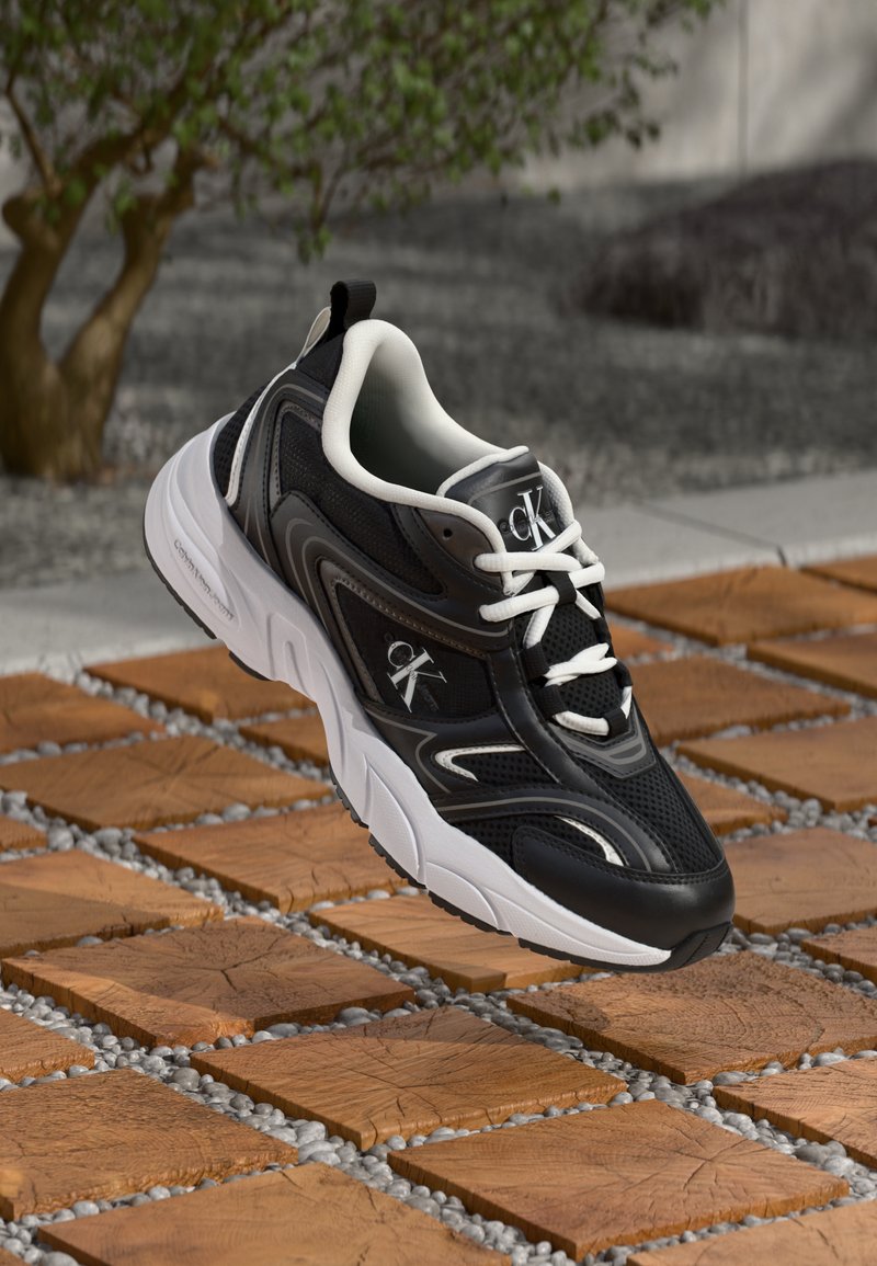 Μαύρα και άσπρα αθλητικά παπούτσια με διχτυωτό και συνθετικό υλικό, με στρογγυλή μύτη, έντονα κορδόνια και λογότυπο στο πλάι.