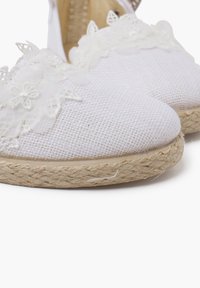 Des espadrilles blanches avec des dessus en tissu texturé, ornées d'accents en dentelle florale et d'une semelle en corde de jute naturelle.