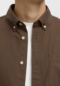 Camisa marrón de botón con collar clásico, con botones blancos, bolsillo en el pecho y una tela texturizada sobre una capa blanca debajo.