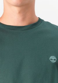 Mørkegrøn bomulds t-shirt med rund hals og et lyseblåt trælogo broderet på venstre side af brystet.