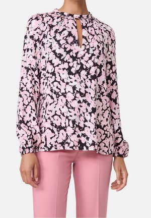 Nainen pukeutuneena pitkähihainen vaaleanpunainen ja musta abstrakti kukkakuosi paita avonaisin pääntiellisin ja vaaleanpunaisiin istuviin housuihin.